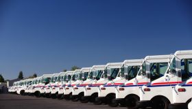 A fleet of U.S. postal trucks sits in a line outside of the USPS center.