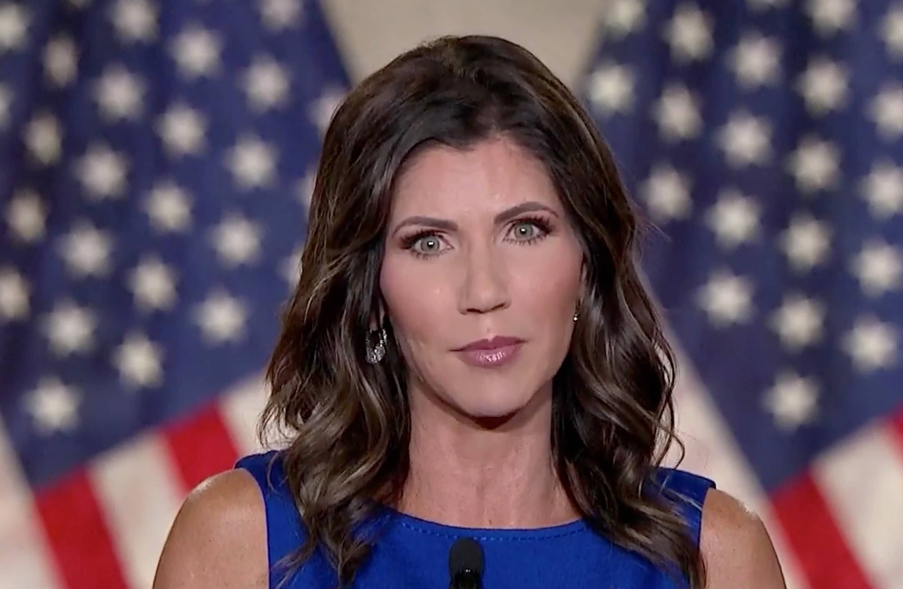, South Dakota Gov. Kristi Noem in a blue dress speaking in front of two american flags