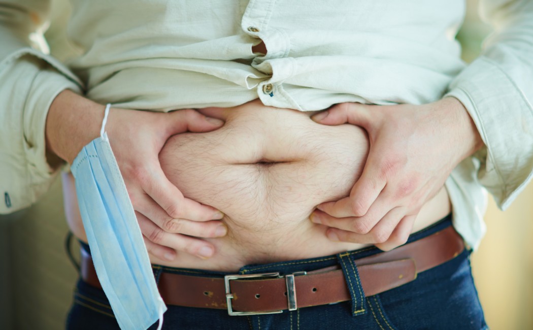 Closeup on unhappy modern 30 years old male in shirt and blue jeans with medical face mask checking body fat by grabbing stomach. Unhappy with weight gain on coronavirus quarantine concept