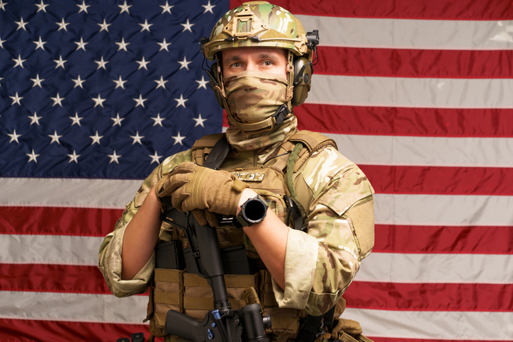 Soldier laid hands on machine gun dressed in ammunition posing on USA flag on background. Army soldier with riffle in camouflage clothing. Flag of United States of America on background