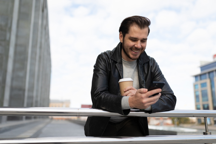 an adult man outside with a cup of coffee in his hands looks at the screen of a mobile phone