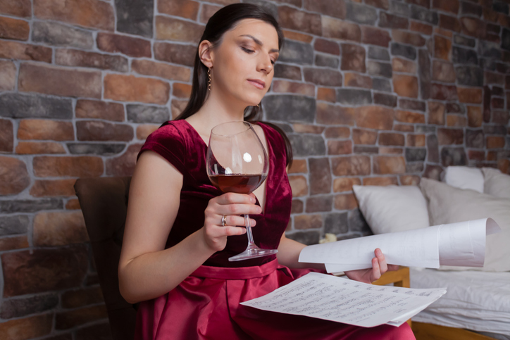 A woman is sitting, looking at musical sheets while holding a glass of wine in her hand.