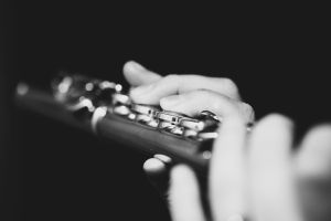 A black and white portrait of the fingers of a hand of a flutist musician gripping down on the valves of a silver metal flute to play a note of a musical piece during a concert.
