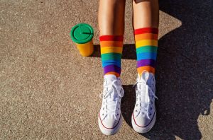 Top view of legs in bright colorful rainbow pride socks at sunlight outdoors. Trendy teen sitting on ground, generation Z lifestyle, pride month.