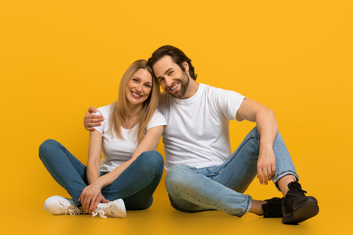 Cheerful young attractive european man with beard and female hugging, sit on floor, isolated on yellow background