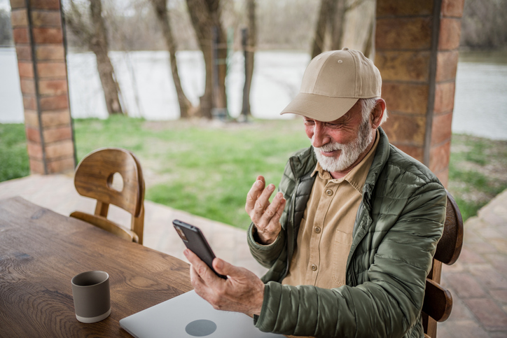 A modern pensioner sits on the terrace of a cottage, surrounded by nature. He is holding a smartphone and is in the middle of a video call with his family