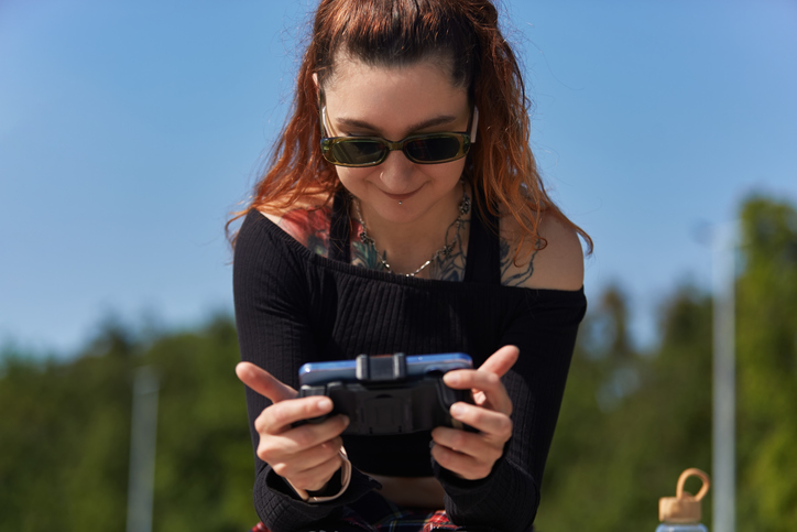 Cheerful gamer playing mobile game on a smart phone with a gamepad. Portrait of a tattooed young adult female in sunglasses gaming online with a modern gadget