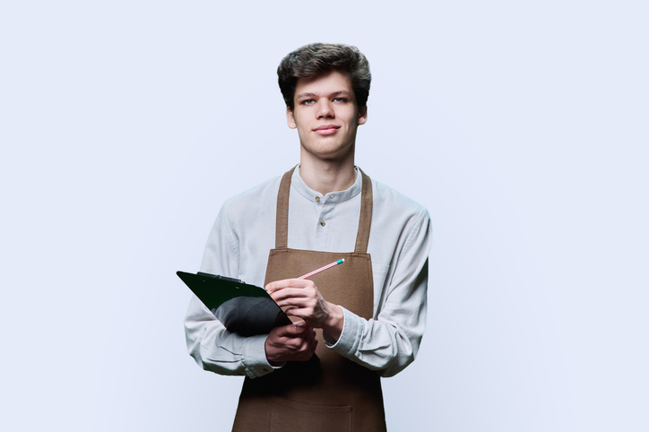 Young guy in apron making notes on clipboard, on white background