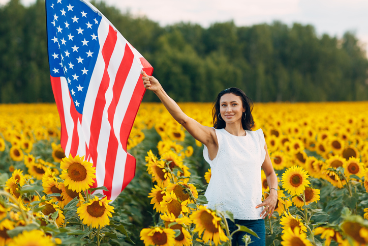 Young woman with American flag in the sunflower field. 4th of July Independence Day USA concept.