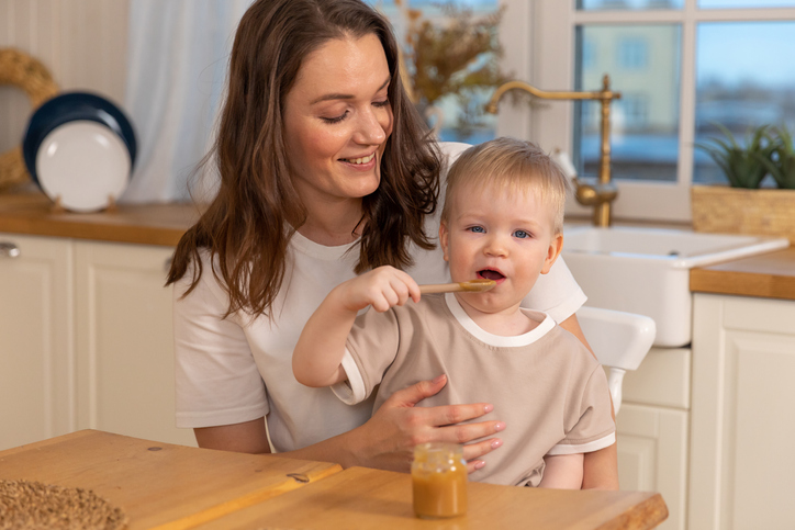 Happy family at home. Mother feeding baby in kitchen. Little boy with messy funny face eats healthy food. Child learns eat by himself holding spoon. Woman mom giving food to kid son. Self feeding.