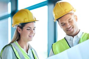 Architect, team and planning with blueprint for construction or architecture with smile for on site project. Man and woman contractors in building floor plan or teamwork strategy with safety helmets