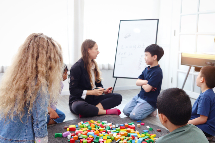 Group of primary school children study and play together in classroom with teacher, diverse students sit on floor while do activities and have fun, kid education, variety and diversity classroom.