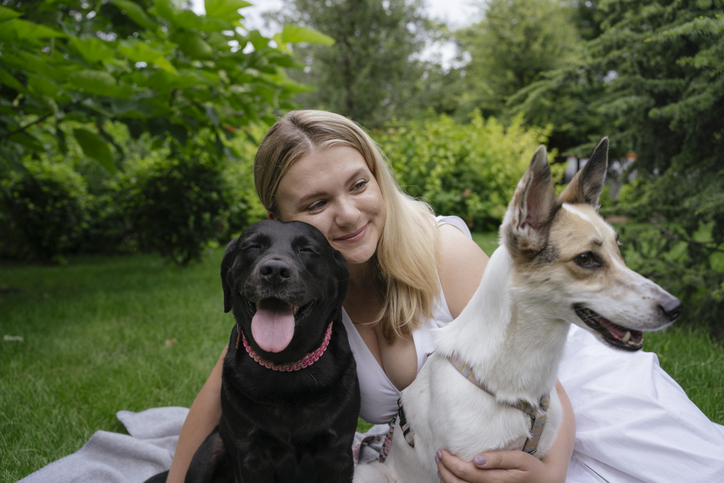 Happy mid adult woman sitting on green grass with two dogs in summer park. Concept of family bonding, pets and togetherness.