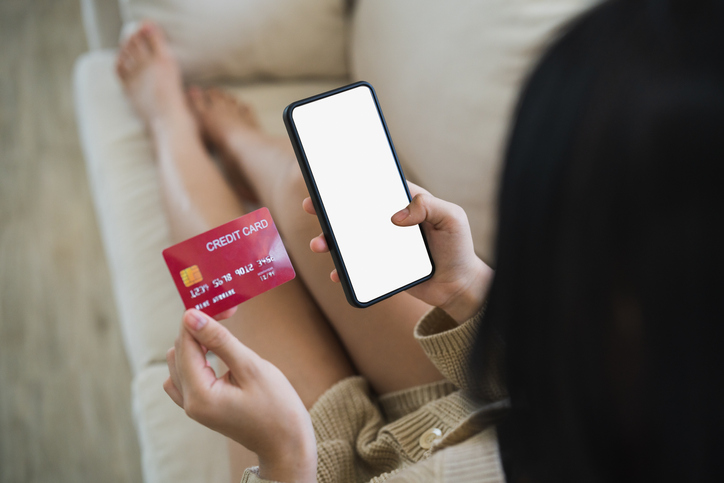 Woman using mobile phone smartphone holding credit card to shopping online. asian woman shopping on sofa couch in living room at home. Online shopping, e-commerce, internet banking, spending money.