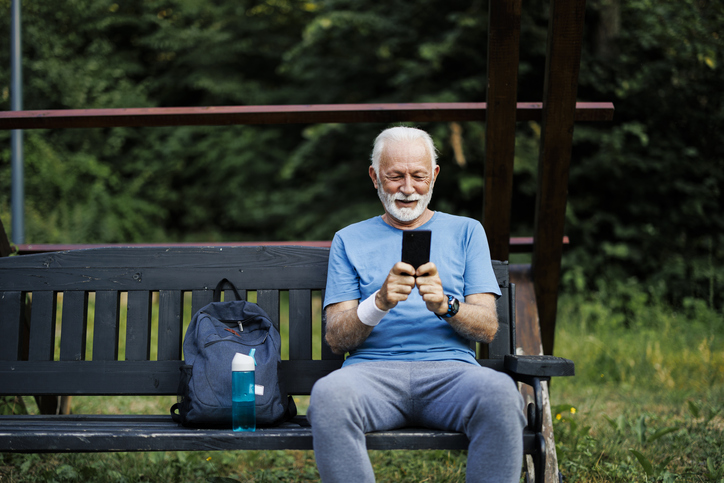 Senior man using smart phone, sitting on bench at park and resting after workout