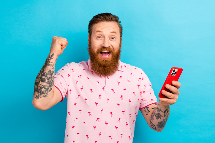 Portrait of overjoyed man with cool tattoo wear flamingo t-shirt hold smartphone clenching fist win bet isolated on blue color background