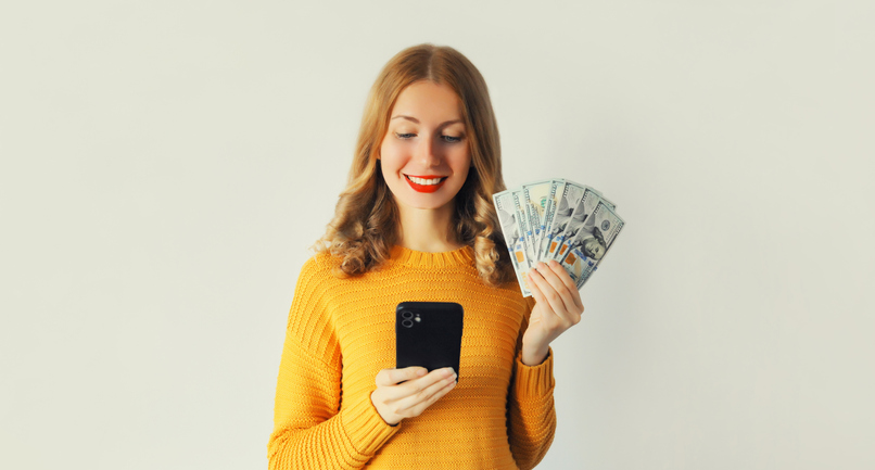 Happy smiling young woman holding phone and cash money in dollar bills