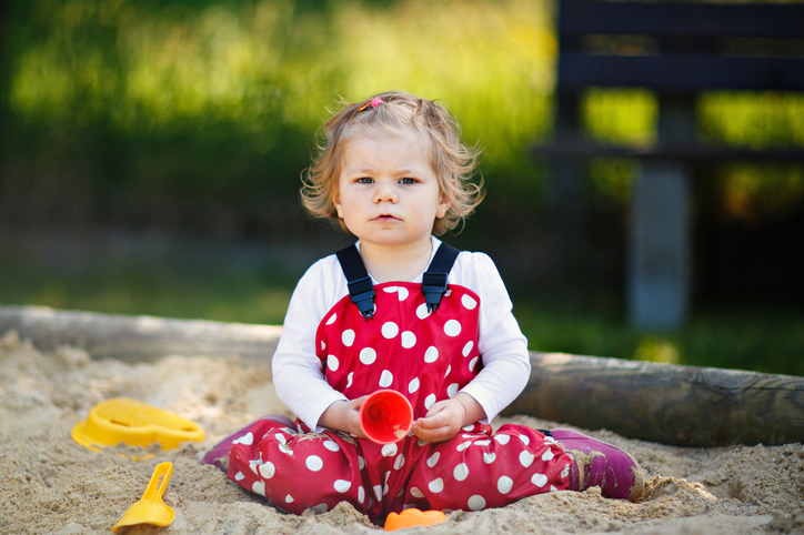 Cute toddler girl playing in sand on outdoor playground. Beautiful baby in red gum trousers having fun on sunny warm summer day. Child with colorful sand toys. Healthy active baby outdoors plays games