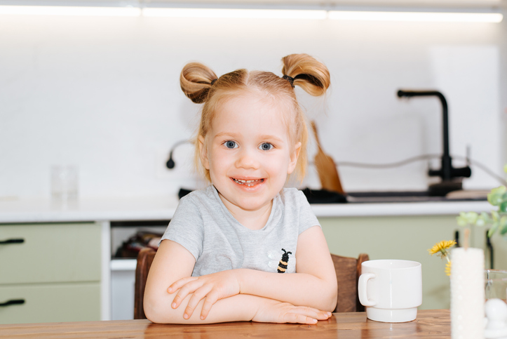Portrait of single cute happy baby girl in kitchen, caucasian smiling kid sitting with mug at table and looking at camera
