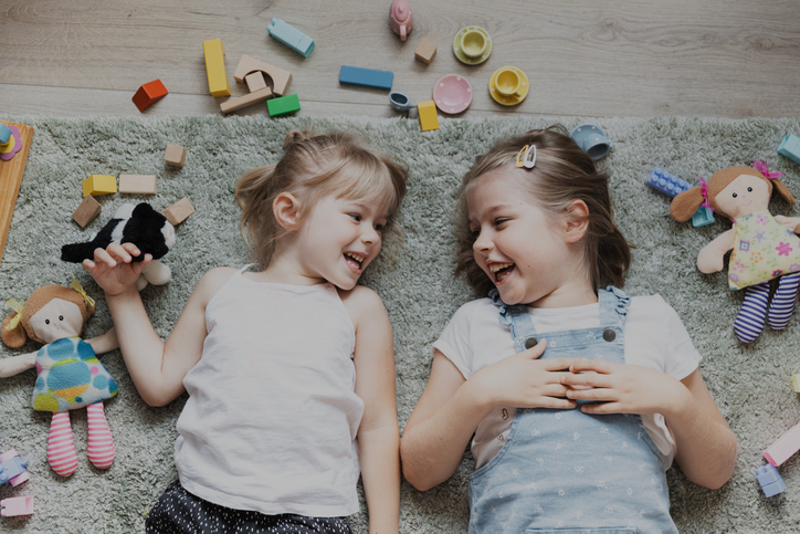 Two small sisters playing with different toys at home, lying on the floor