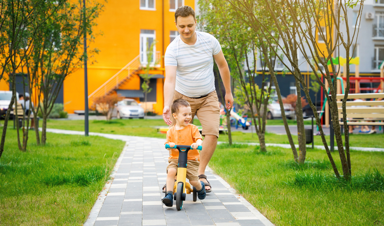 A cute happy toddler boy of two or three years old with dad rides a bicycle or balance bike in a residential complex on a sunny summer day. Happiness spending time with family