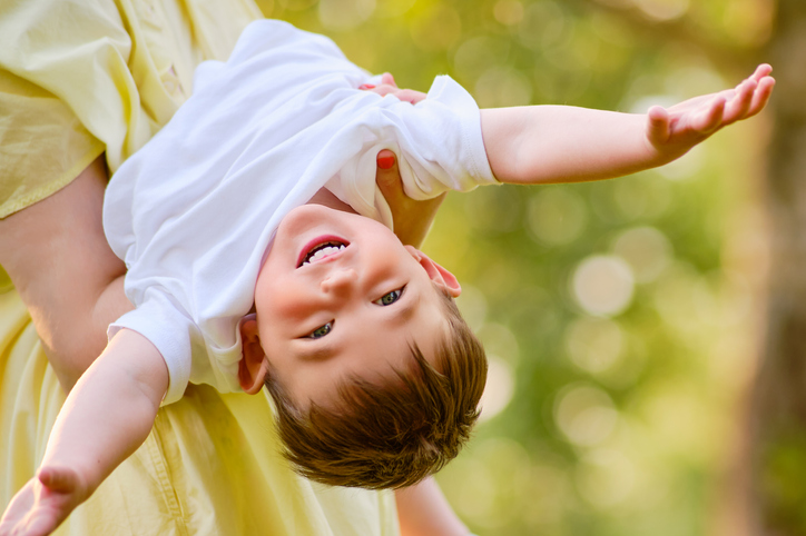 Baby upside down in her mother's arms on a nature walk. Happy child with mom playing on the green grass in the summer park. Kid aged about two years (one year eleven months)