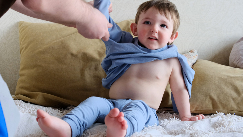 Hand father puts on blue pajamas on the baby boy. Dad man dresses clothes for child on the bed with pillows. Kid about two years old (age one year nine months)