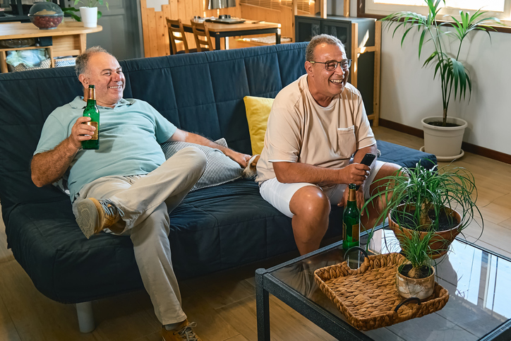 Two male friends sitting on couch watching tv, football game or streaming program. Middle aged oversize men spending leisure time at home together in front of television.