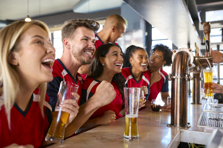 Multi-Cultural Group Of Friends Wearing Team Shirts In Sports Bar Celebrating Watching Game On TV