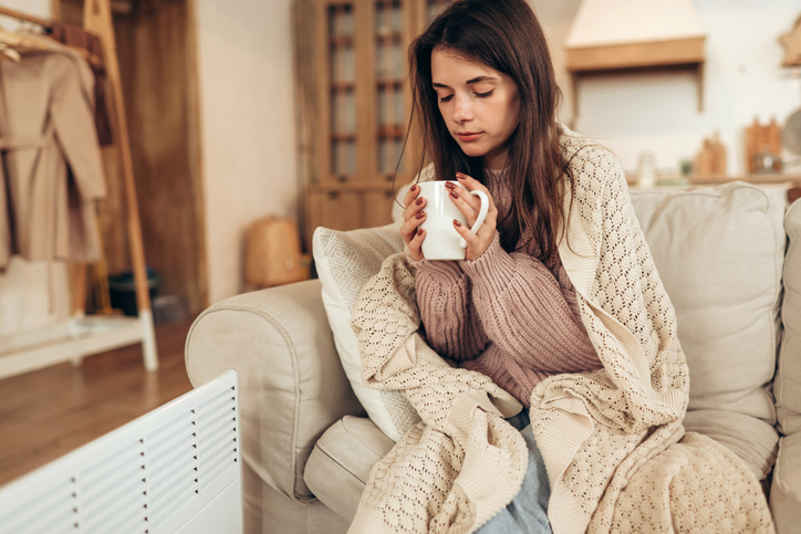 Woman freezes in winter time. Young woman with plaid near electric heater, holding a cup of hot drink while sitting on sofa at home. Keep warm.