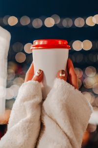 Female hands holding a plastic cup with hot tea or coffee against the background of bokeh garland lights.