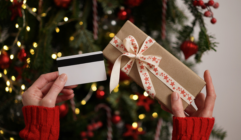 Hands of a woman in a red sweater holding a credit card and a gift box on the background of a Christmas tree close-up. Christmas and New Year shopping online, credit card payment. Buying gifts online.