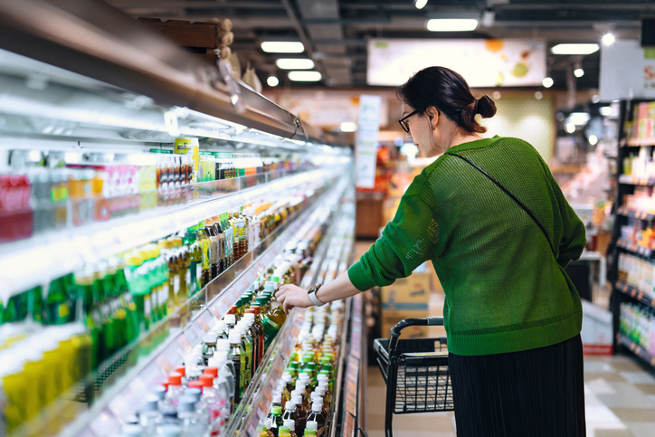Young Asian woman with shopping cart picking up a bottle of drink over the refrigerated shelves in supermarket. Routine grocery shopping. Making healthier choice. Smart eating. Concept of choice and customer awareness