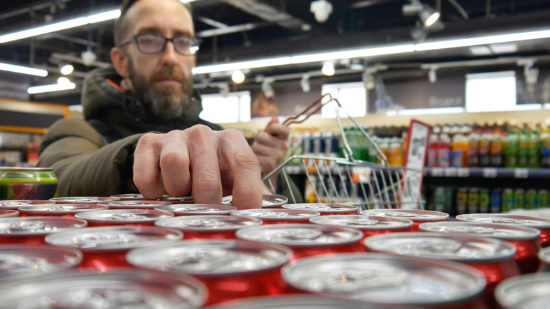 Close-up of many red cans of cola in a supermarket and a buyer with a shopping basket taking one