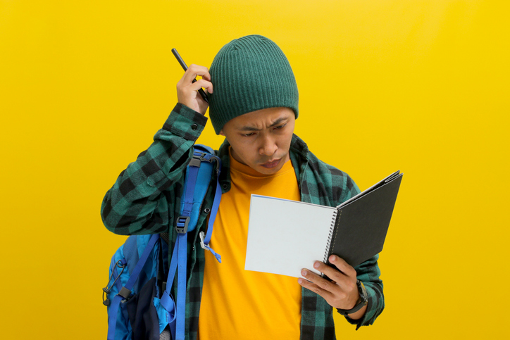 A confused young Asian student scratches his head while reading a book
