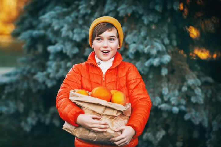 A Boy In A Bright Orange Jacket And A Yellow Hat Holds A Large Package With Oranges