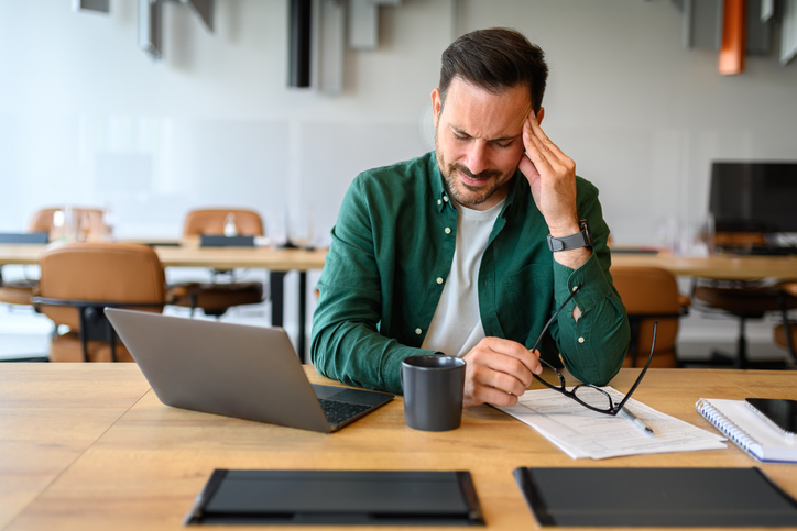 Young businessman holding eyeglasses and touching head in pain while working over laptop in office