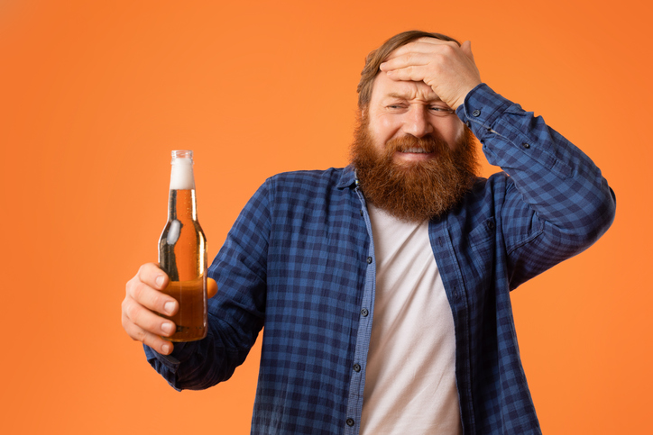 unhappy redhaired bearded man holding beer bottle touching head, studio