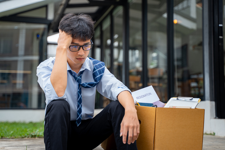 Sad young businessman sitting at a curve side road with a box full of his office stuff beside him