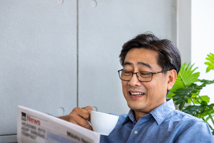 Senior Asian man smiling while reading newspaper and drinking coffee at home