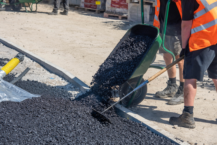 Construction Workers Using Wheelbarrow to Spread Asphalt on Sunny Day at Roadwork Site