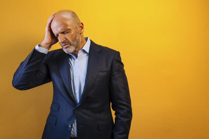 a man in a suit with his hands on his head looking distressed. He is against a yellow background.