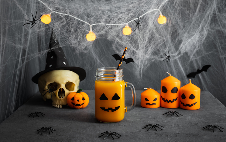 Halloween juice in a glass jar with jack lantern face, skull, candles, spiders and cobwebs on gray background. Halloween drinks table setting.