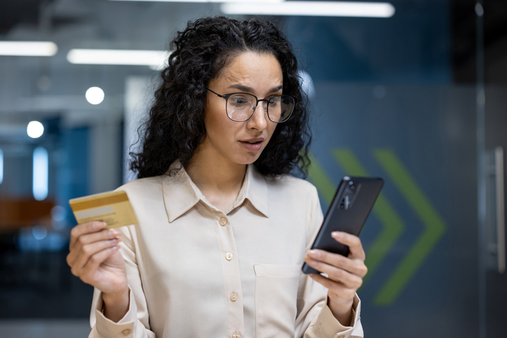 Worried woman using phone and holding credit card in office setting, analyzing online transaction, concerned about financial security, digital payment risks, experiencing online banking issues