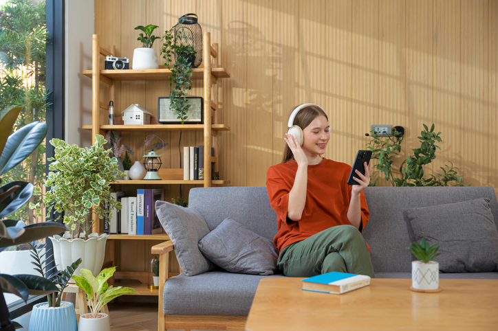 Young woman listening to music on wireless headphones over a mobile app while sitting on sofa in living room at home.