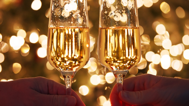 Holiday dynamic postcard. Two glasses with champagne wine and lighted fireworks on a glitter background with bright gold lights of a night city. Meeting the new year. Christmas party