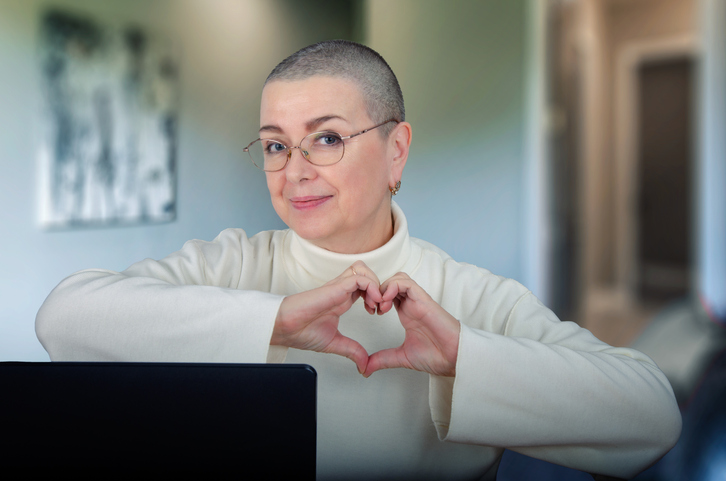 Elderly Woman with Shaved Head and Glasses Displays Love and Resilience, Making Heart Shape with Hands While Working.