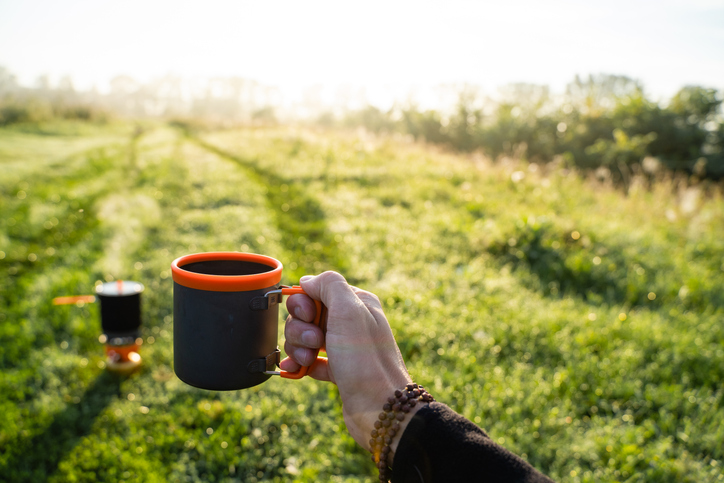 A person is standing in a lush green field, enjoying a steaming cup of coffee, holding a cup of drinkware in their hands amidst the vibrant grass and beautiful landscape
