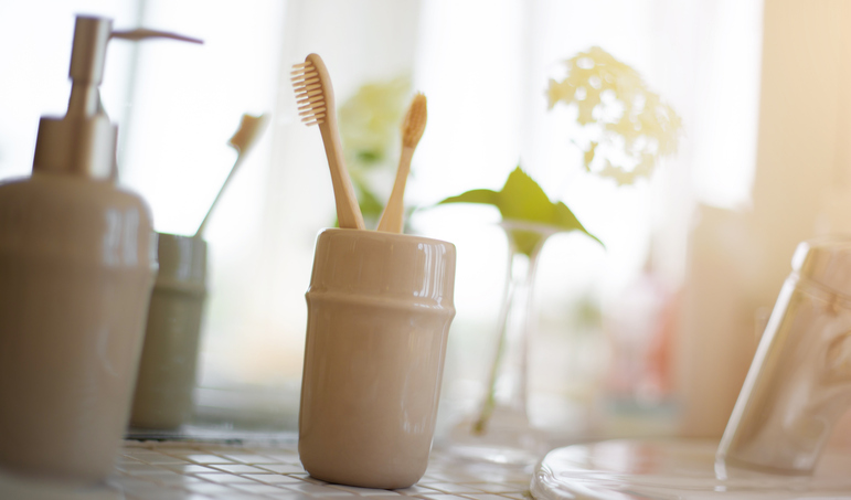 Close-up of a bathroom countertop featuring a ceramic holder with wooden toothbrushes, a soap dispenser, and a vase with flowers in the background. The soft lighting creates a calm, fresh atmosphere