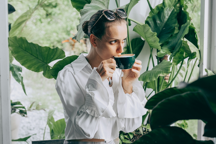 Woman enjoying coffee amidst lush greenery in a serene indoor garden setting.